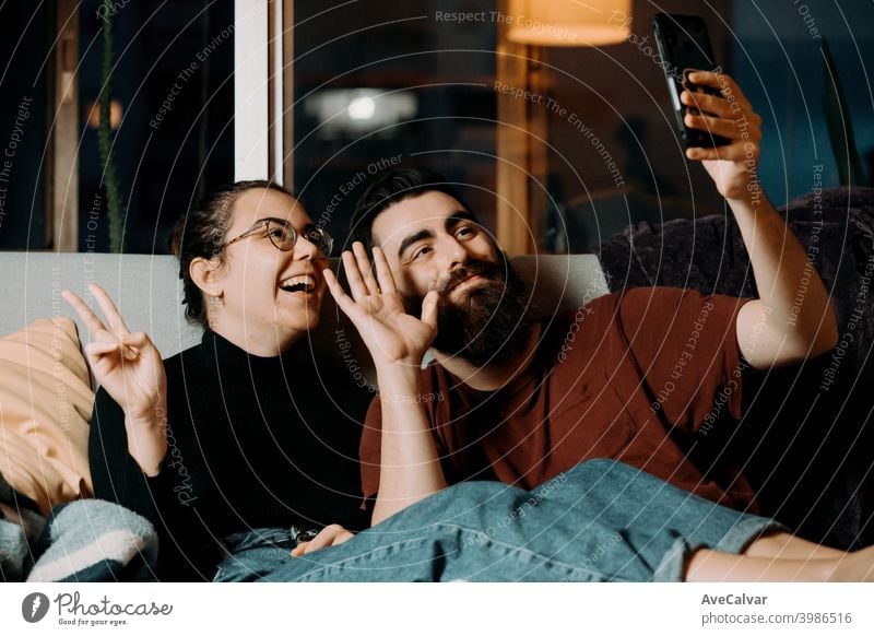 A young couple on the couch greeting to the mobile phone and smiling while on pandemic affectionate embracing hugging humor wife husband photograph ethnicity