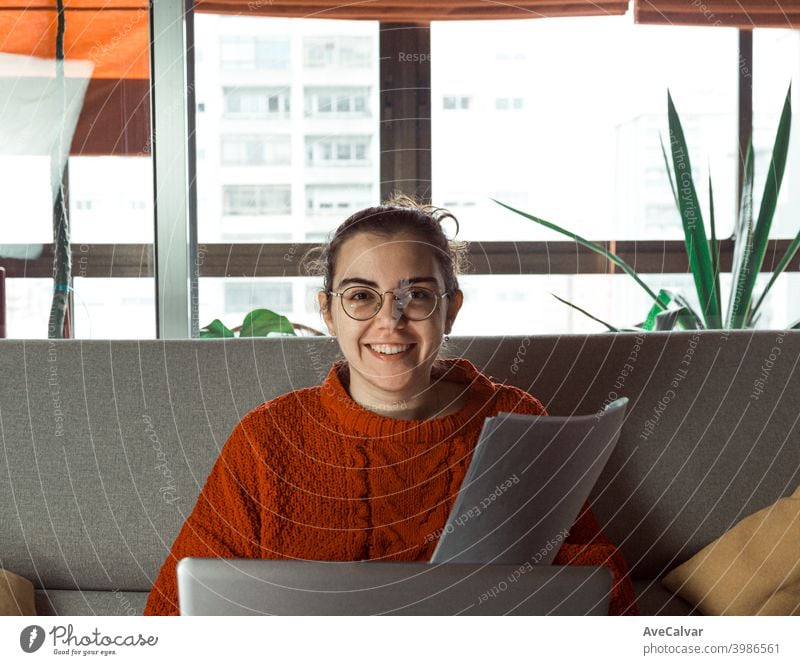 A young woman on glasses working on his laptop on the couch at home while smiling to camera documents horizontal job living relaxed research successful teacher