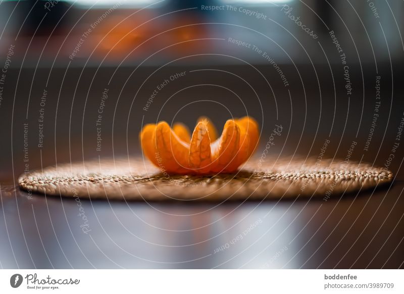 A peeled and unfolded clementine lies on a coaster made of rice straw. Photographed against the light from the height of the tabletop, focused on the clementine.