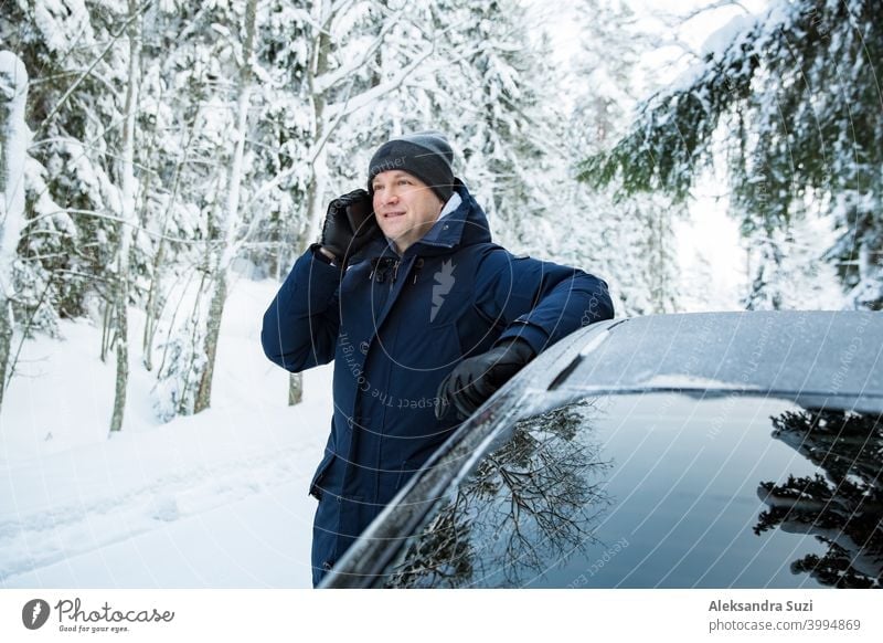Man in warm winter clothes standing at the car, using phone. Snowy winter country road, car covered with ice, Beautiful forest under the snow. help call