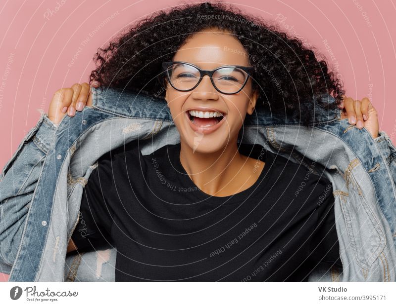 Isolated shot of curly cheerful woman tries on new denim jacket, wears optical glasses, smiles broadly, has perfect mood, poses against pink wall. Positive emotions. Afro female in stylish clothes