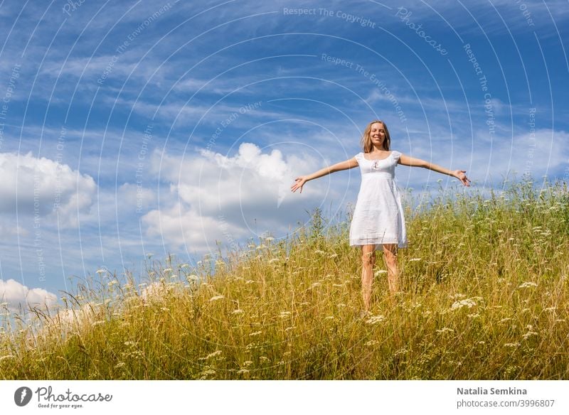 Young girl in white dress is standing on grass hill on blue sky background in summer day. young 20s real people meadow freedom outdoor hands landscape