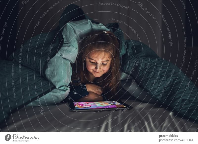 Little girl having fun watching, playing and listening to stories on tablet computer laying under duvet night bedtime child home bedroom read kid relaxation