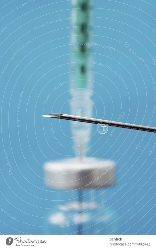 A drop of a vaccine hangs from a cannula and shows the reduced image of a small glass bottle with septum and syringe Macro with shallow depth of field and blue background