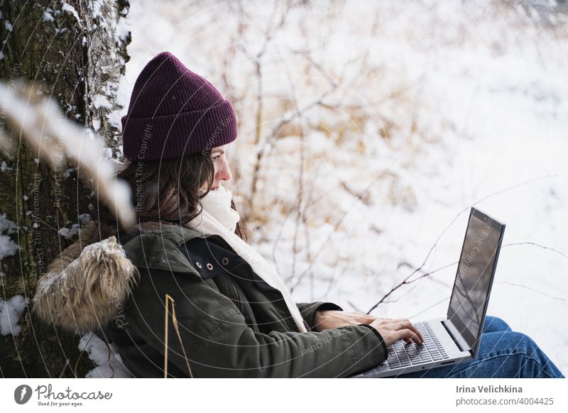 Young girl, woman working in laptop. Walking in beautiful winter forest among trees, firs, covered with snow. Magnificent nature and views. Fashionable image, clothes, parka, hat, mittens, blue jeans