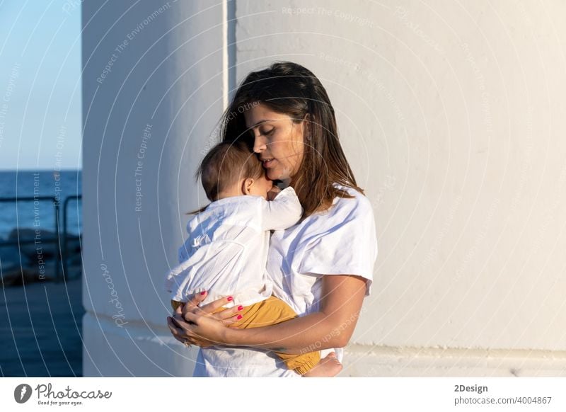 Young mother holding her little baby against white wall in sunny day child newborn love family woman care motherhood young childhood cute person happy girl