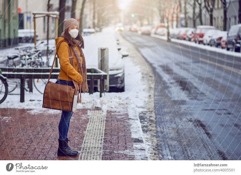 Midde-aged brunette woman standing at a bus stop, wearing a protective face mask due to corona virus, waiting for her bus to bring her to work middle-aged