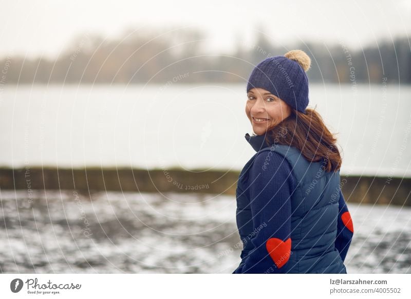 Happy middle-aged brunette woman in sweater with knit hat turning and looking at the camera face portrait winter smiling love copy space water lake cold