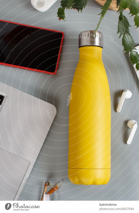 Yellow insulated bottle on grey desk surrounded by modern gadgets and plant yellow inculated reusable mockup vase top view earphones smartphone ecologic water