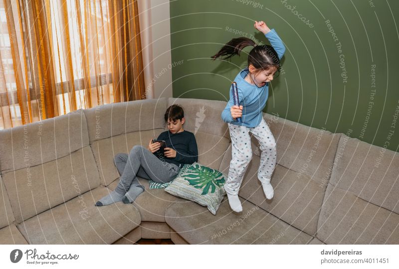 Girl jumping and playing on the sofa while his brother looks at the tablet sitting girl couch homewear teen fun shout caucasian young people lifestyle cute