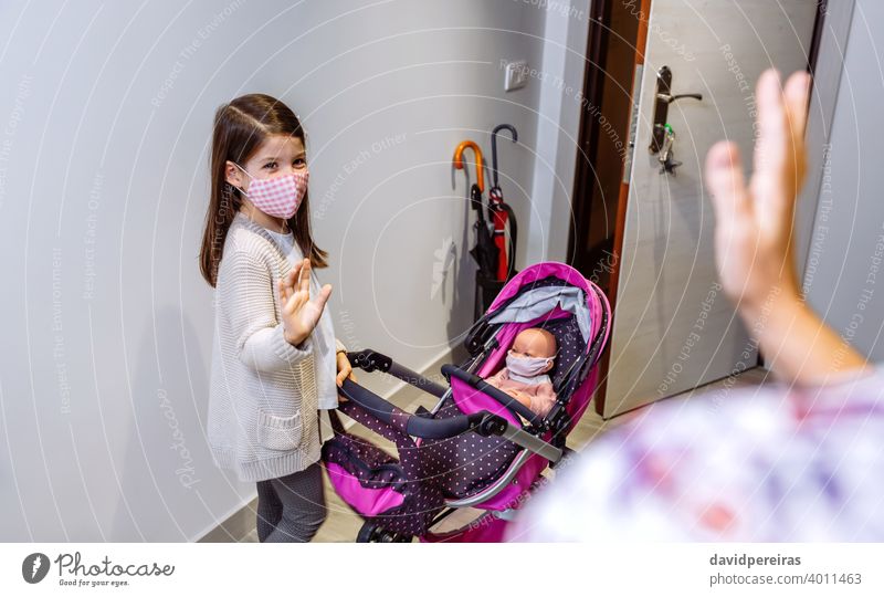 Girl with mask ready to go for a walk with her doll with mask saying goodbye to his mother girl coronavirus protective mask stroller toy waving ready to go out