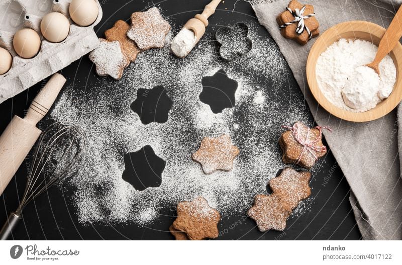 Star shaped baked gingerbread cookies sprinkled with powdered sugar on a black table flour ingredient napkin egg plate spoon rolling pin icing handmade bakery