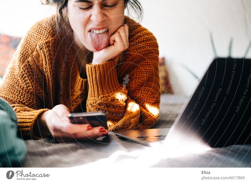 A woman lies at home in bed next to her notebook and sticks her tongue out at her smartphone. Annoyed by the home office. Bed Home Office laptop Notebook Woman