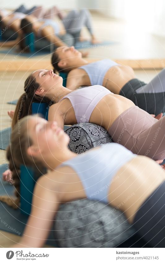 Restorative yoga with a bolster. Group of three young sporty attractive women in yoga studio, lying on bolster cushion, stretching and relaxing during restorative yoga. Healthy active lifestyle