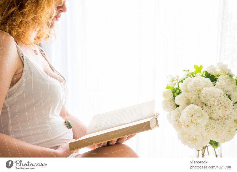 Pregnant woman touching her belly while she is readind a book pregnant bed pregnancy reading redhead red hair morning young girl window freckles sunday relaxing