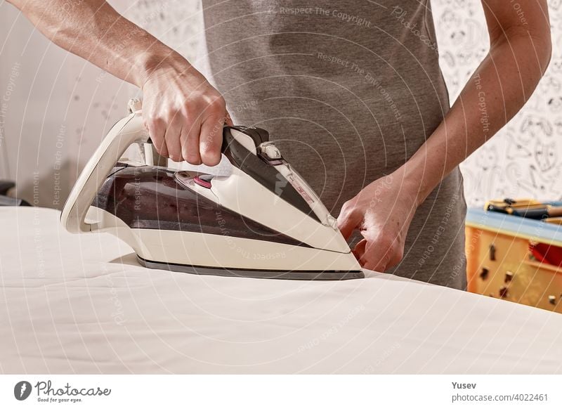 Hands of a caucasian woman holding an iron. Woman is ironing clothes on an ironing-board. Housework. Close-up. hands ironing board housework close-up grey