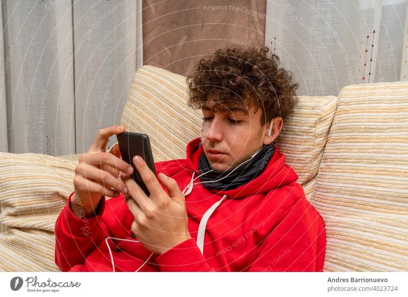 Young attractive man with curly hair dressed in a red sweatshirt using mobile in living room at home wearing a mask to protect himself from coronavirus