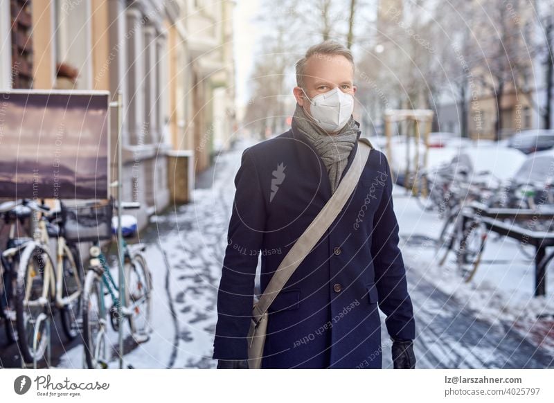 Man wearing a protective surgical face mask during the Covid-19 or coronavirus pandemic and winter overcoat with leather bag over his shoulder walking down a snowy urban street with parked bicycles in close up looking aside