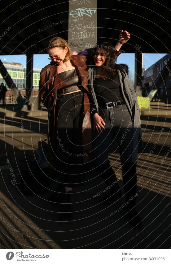 Lara and Estila Woman Long-haired Stand Cool Friendship Coat Easygoing Blonde Dark-haired Architecture Bridge sunny Sunlight togetherness at the same time fun