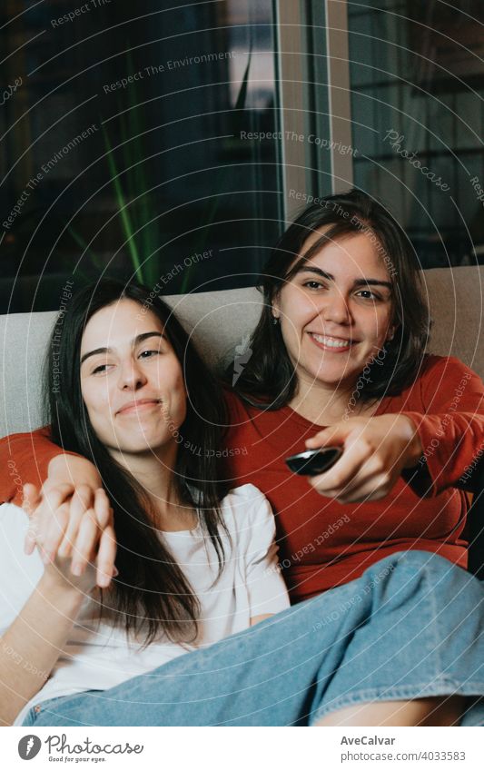 A lovely couple of two women looking the Tv in a modern flat on the couch while smiling affectionate answering cuddling embracing friends girls headshot