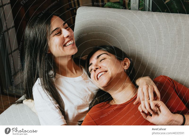 A lesbian couple smiling and laughing having fun on the couch of a modern flat while hugging each other with love affectionate answering cuddling embracing
