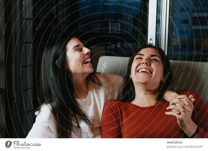 A couple of woman laughing on the couch during a happy night in the flat of the city affectionate answering cuddling embracing friends girls headshot horizontal