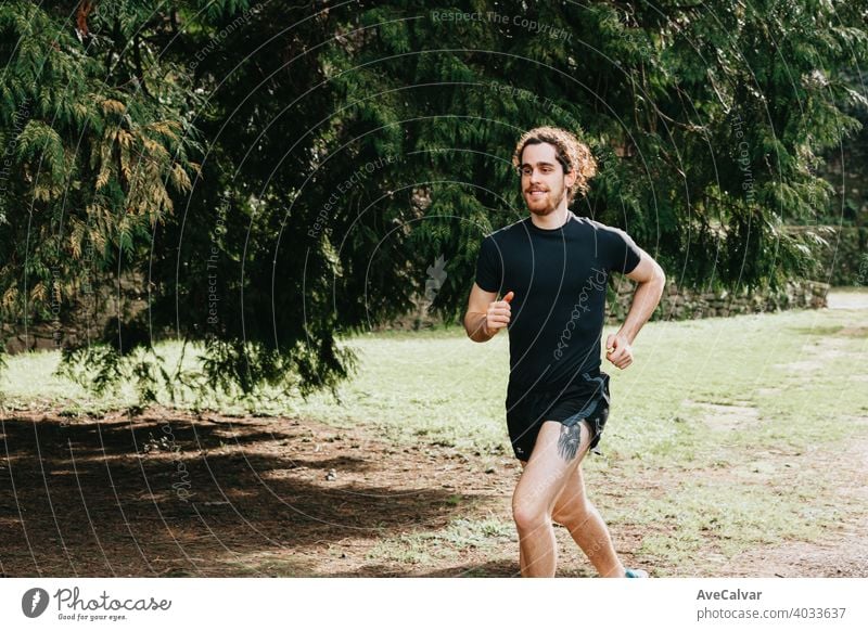 A long hair male running between the trees during a sunny day in the park with copy space 20s exercising horizontal marathon runner sprinting touching jogging