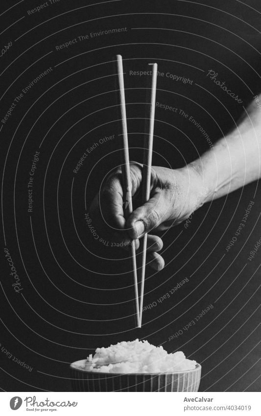 Minimalistic old hands grabbing japanese chopsticks over a bowl of rice concept shot on cinematic tones over a black background in black and white minimal
