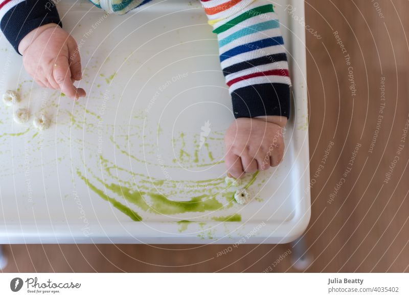 Young baby in rainbow stripe outfit spreading puff cereal and green puree on high chair tray; baby led weaning pincer palmar infant child 6 months old