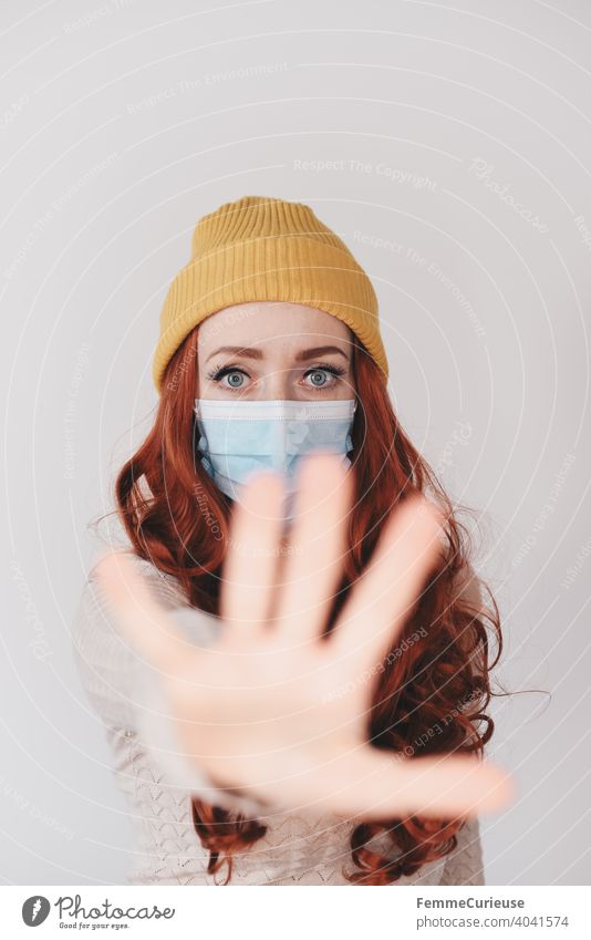 Young redheaded woman with yellow hipster beanie cap , medical protection mask and outstretched hand towards camera as sign for please keep distance gap