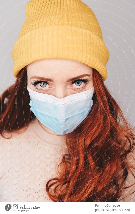 Young red-haired woman with yellow hipster beanie cap and medical protective mask looks into camera with penetrating look (portrait) haunting Looking