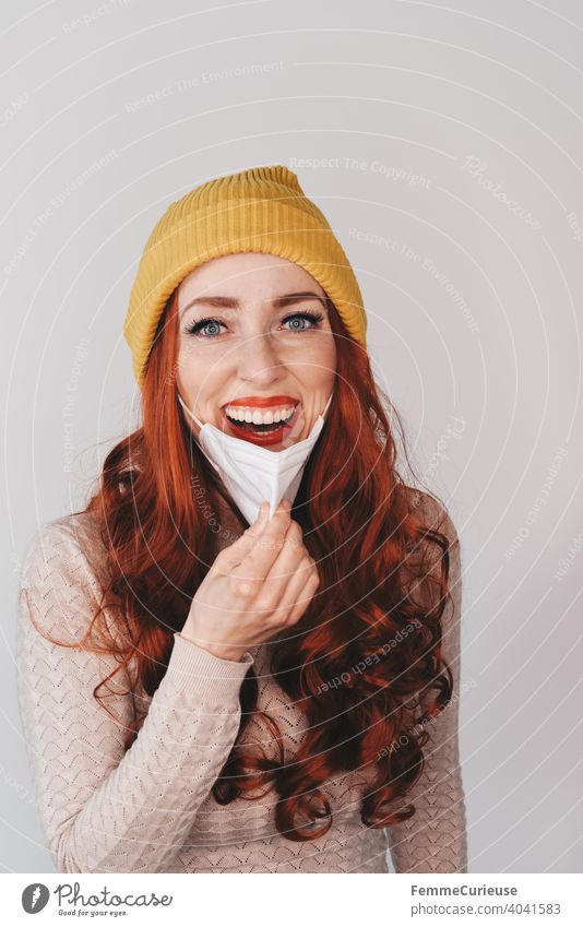 Young redhead smiling woman in yellow hipster beanie cap pulls FFP2 mask away from face below Woman Young woman Beanie hat Yellow Red-haired Curly Sweater