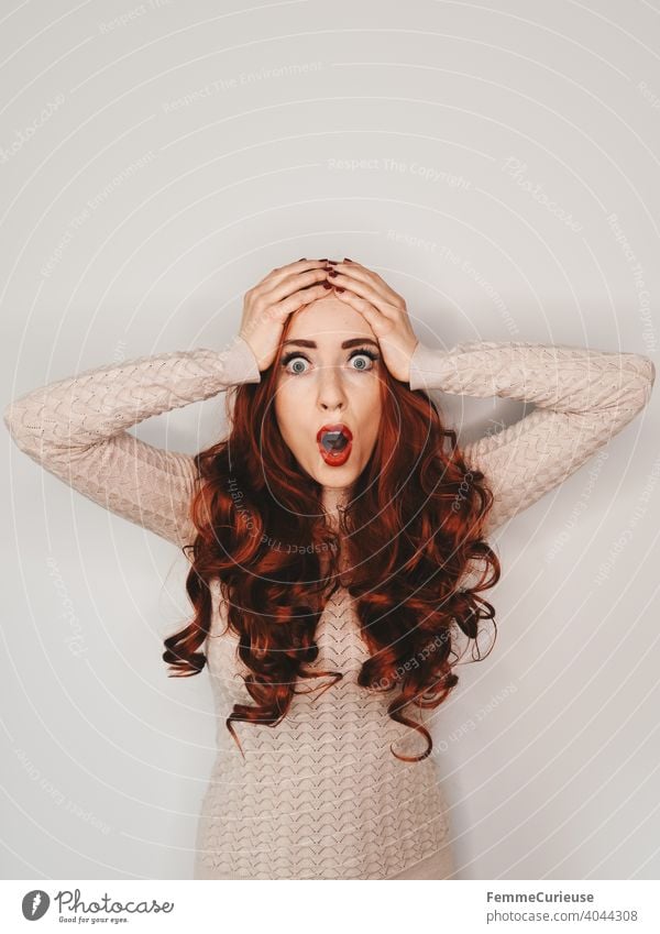 Woman with long red curly hair and frightened expression slaps both hands together over her head shocked astonished Neutral background Sweater skin-coloured