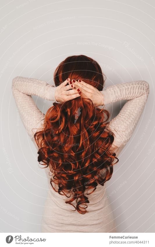 Back view of a woman with long red curly hair in a skin-colored sweater holding her hair together with both hands stick together stop To hold on Nail polish Red