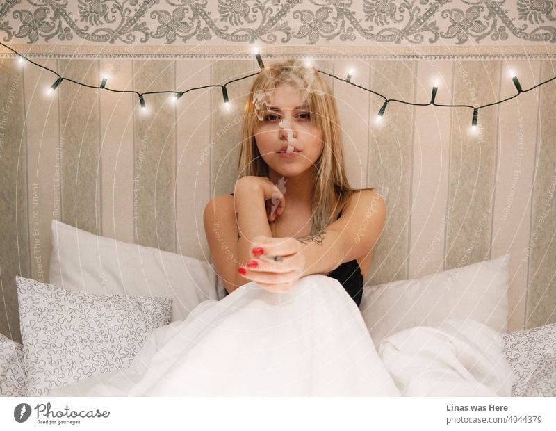 A gorgeous blonde girl is smoking a cigarette in bed. Sheets and pillows are white, the lighting is warm, and the wallpaper behind this beauty is vintage. A pretty woman enjoys her guilty pleasure.