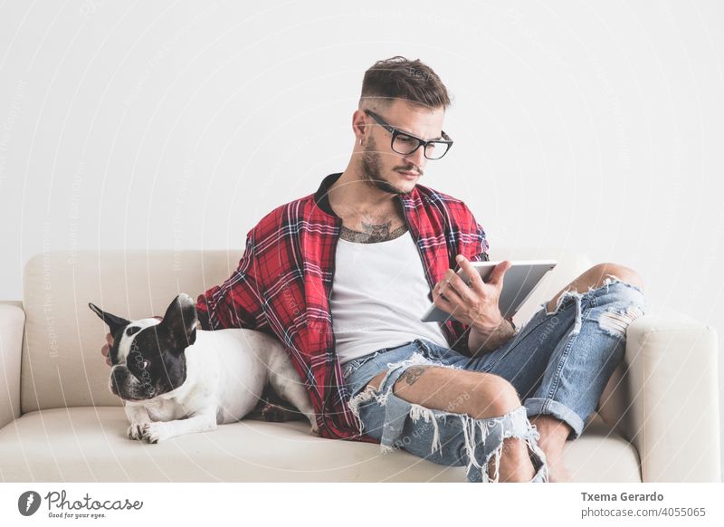 Young hipster looking at his tablet next to his dog sitting on the sofa french bulldog boy piercing pierced hat earrings shirt beard pet puppy animal young man