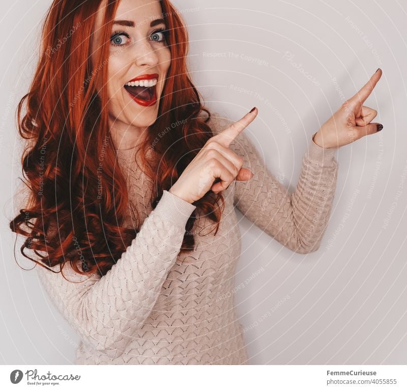Woman with long red curly hair points with both hands and index fingers at something while laughing into the camera Clue indicative Indicate Forefinger