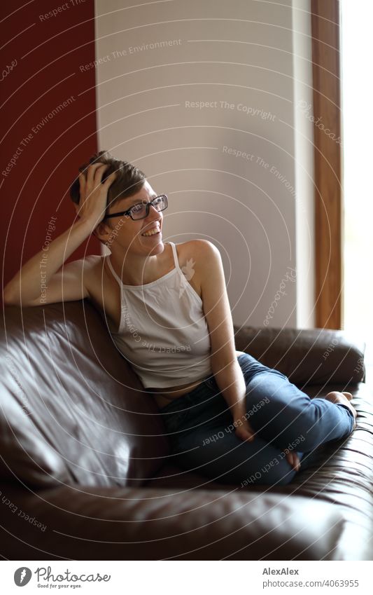 Young woman in jeans and top with glasses sits on a brown leather couch and laughs Woman Slim Top Eyeglasses Shoulders Skin Barefoot Large Athletic Articulated