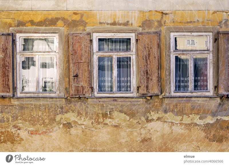 The three old wooden windows with their open shutters, the curtains concealing the view and the peeling plaster all around reflect the opposite side of the street.