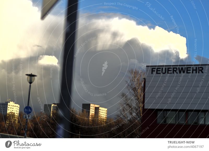 FEUERWEHR is written in bold letters on the wall of a building, inconspicuous nonetheless because the late sunlight makes clouds and prefabricated buildings in the background shine brightly, while lantern, sign and tree make everything even more confusing