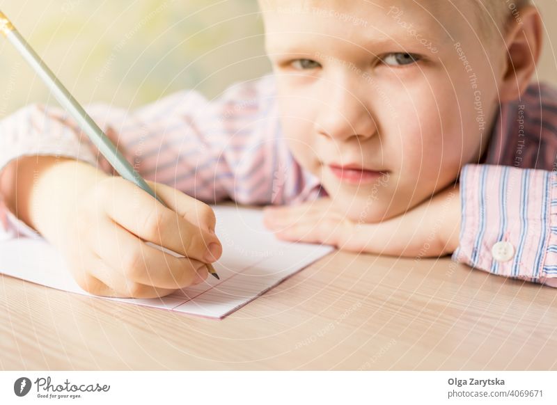 Cute  boy writing his homework and looking at the camera. kid study doing cute concentrated blond pink shirt pencil copybook notebook school face one day hand