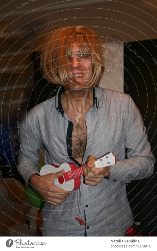 Cheeful man in grey unbuttoned shirt and in blonde wig with toy guitar in his hands portraying singer or musician in room at home party. Birthday celebration at home. Party at home. Waist portrait.