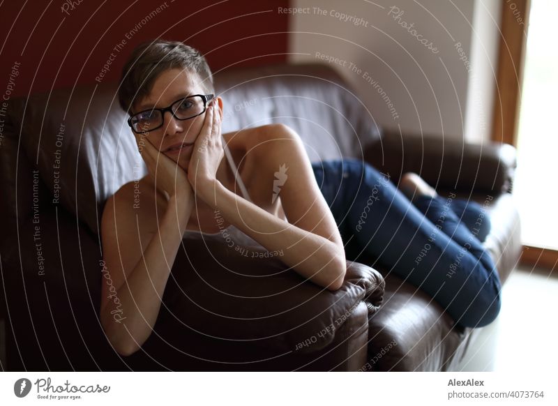 Portrait of a young woman on a brown leather couch Woman Young woman Slim Top Eyeglasses sits Skin Large Athletic Articulated inside Room studio Bright daylight