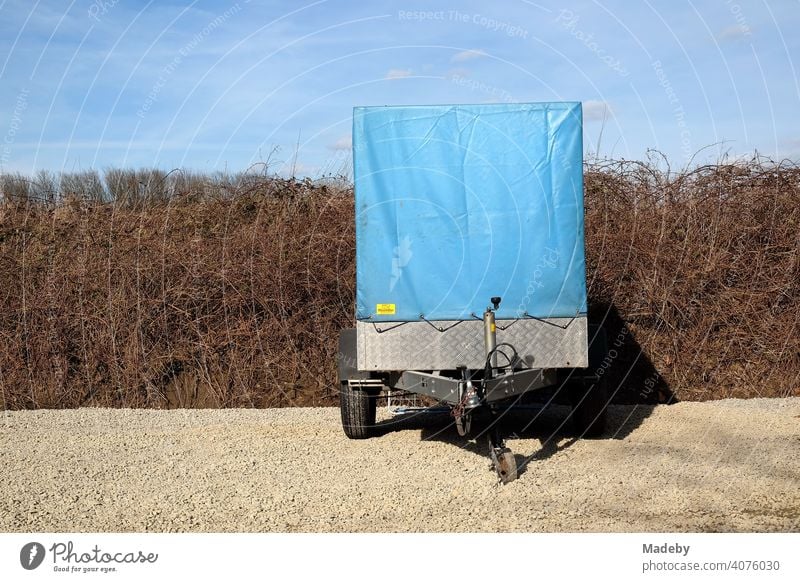 Car trailer with high blue tarpaulin made of plastic in front of brittle hedge and blue sky on gravel in natural colors in Lemgo near Detmold in East Westphalia-Lippe