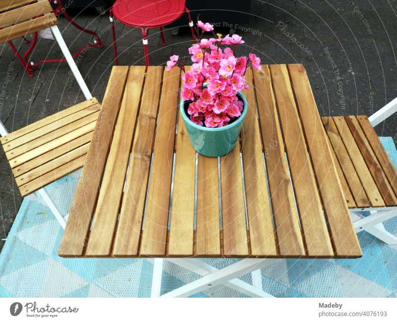 garden table made of light wood with bistro chairs with flower pot with plant with pink blossoms in front of a shop in the old town of Detmold in East Westphalia-Lippe