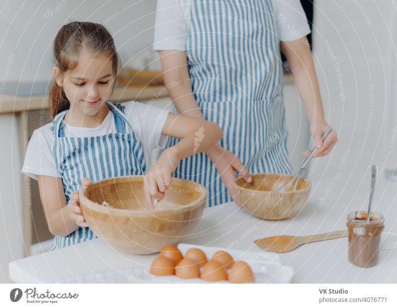 Mother gives culinary lesson to little child, stand next to each other, mix ingredient in big wooden bowls, make dough together, use eggs, flour and other products. Family, cooking, motherhood
