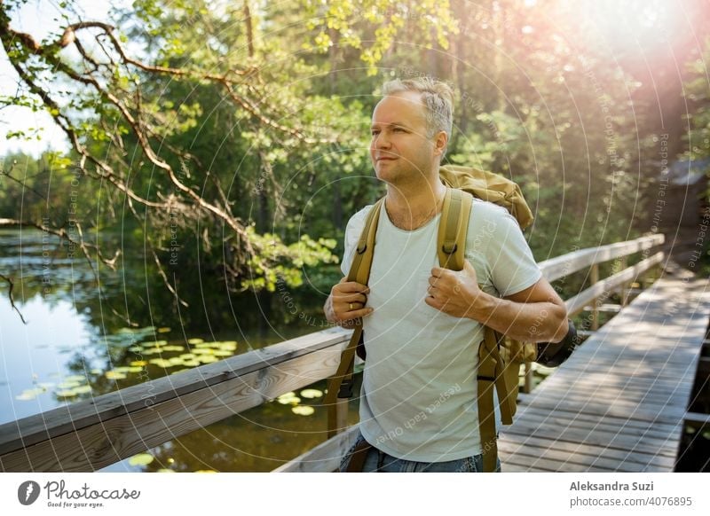 Mature man exploring Finnish nature in summer, walking across the bridge. Hiker with big backpack traveling in forests. Summer Scandinavian landscape of lakes and woods.