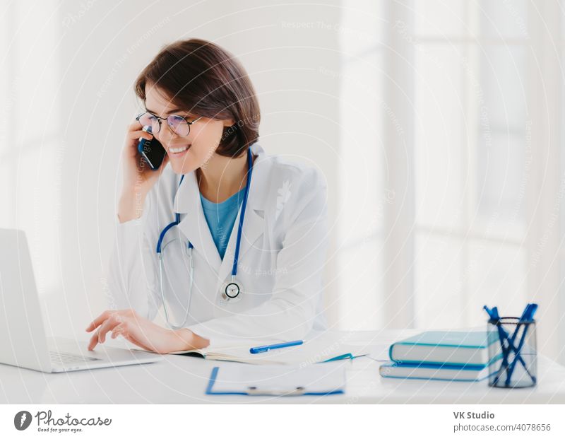 Online consultation concept. Cheerful female medical worker talks via cellular, searches useful information in internet, works on laptop computer, sits at desktop with notebooks, clipboard, pens