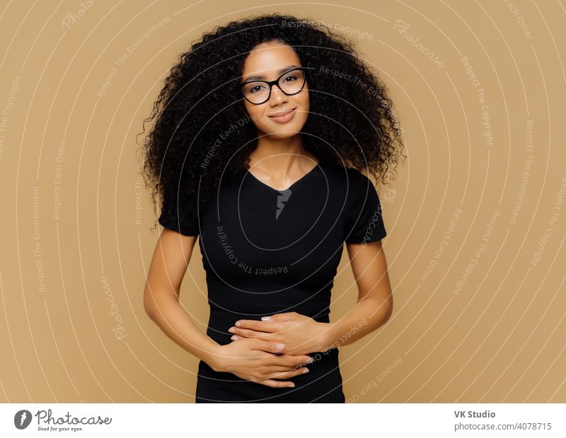 Pleased Afro American woman with curly hair, keeps hands on belly, feels good after eating nutritious delicious dinner, looks happily at camera. Young delighted female finds out about pregnancy.