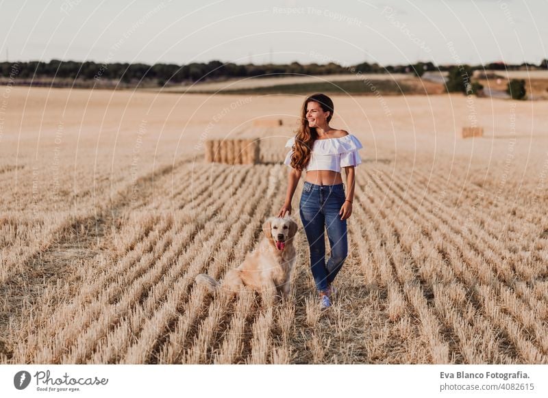 young beautiful woman walking with her golden retriever dog on a yellow field at sunset. Nature and lifestyle outdoors shine relaxation summer fashion harmony